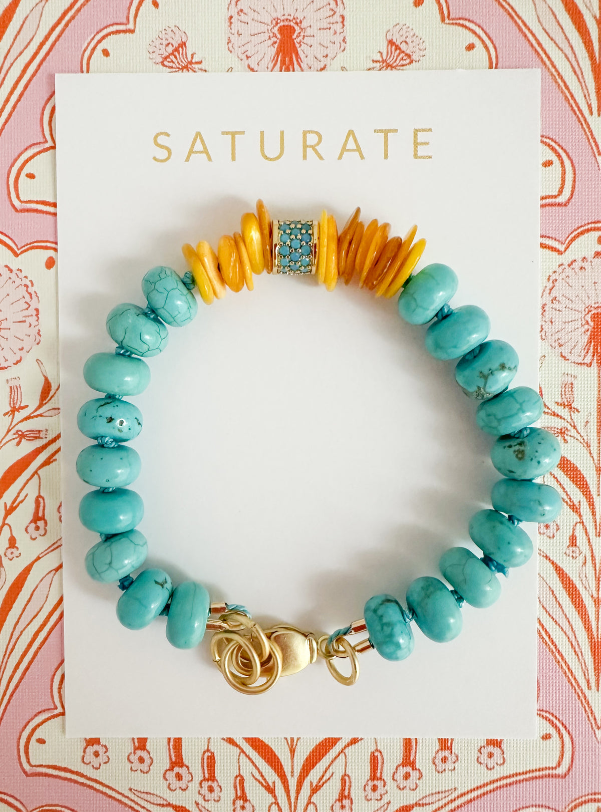 A turquoise and orange beaded bracelet with a gold clasp, displayed on a white Saturate brand card against a pink and orange patterned background.