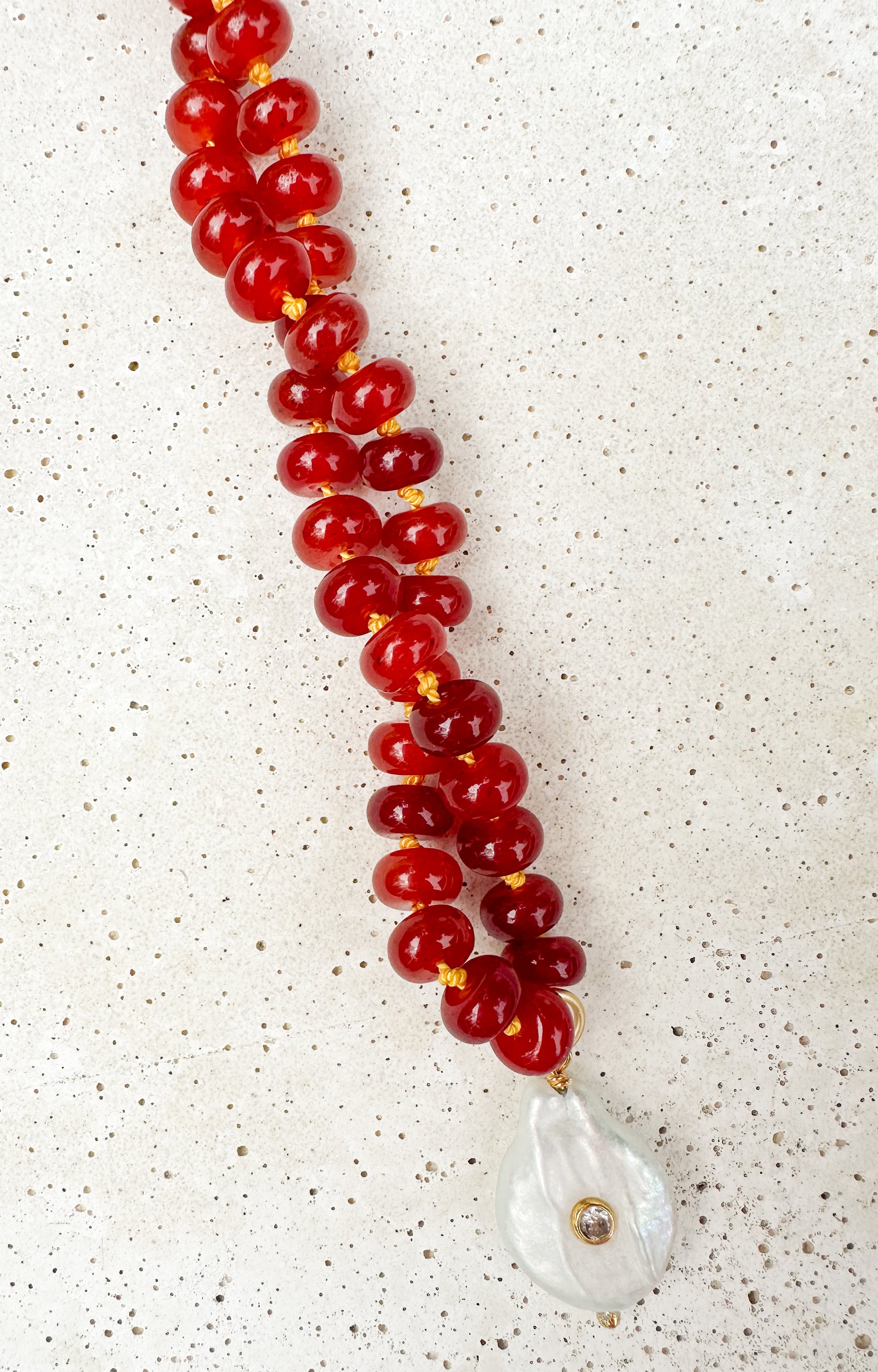 A beaded necklace featuring glossy red round beads strung on gold-toned cords, with a teardrop-shaped white shell pendant accented by a small gold bead, displayed on a textured light beige surface.