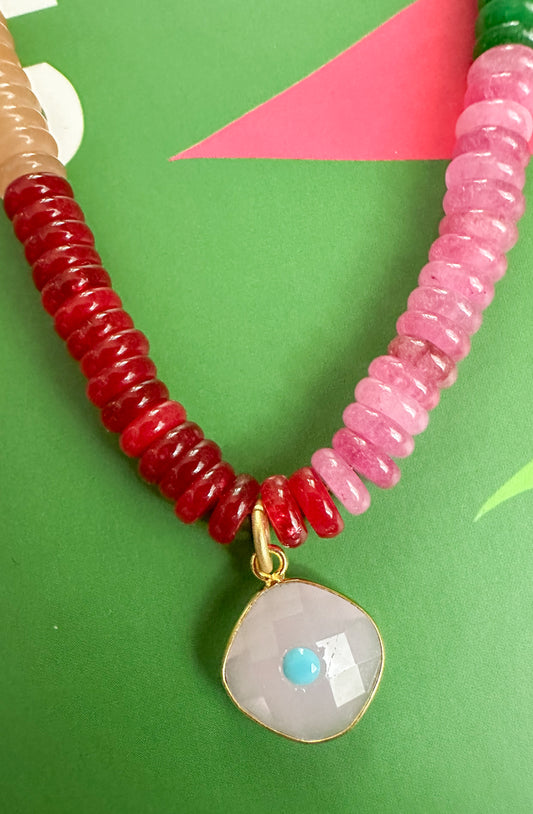 A beaded necklace with a split design of red and pink beads, featuring a central faceted white stone pendant with a small blue center and gold trim, displayed on a green background.