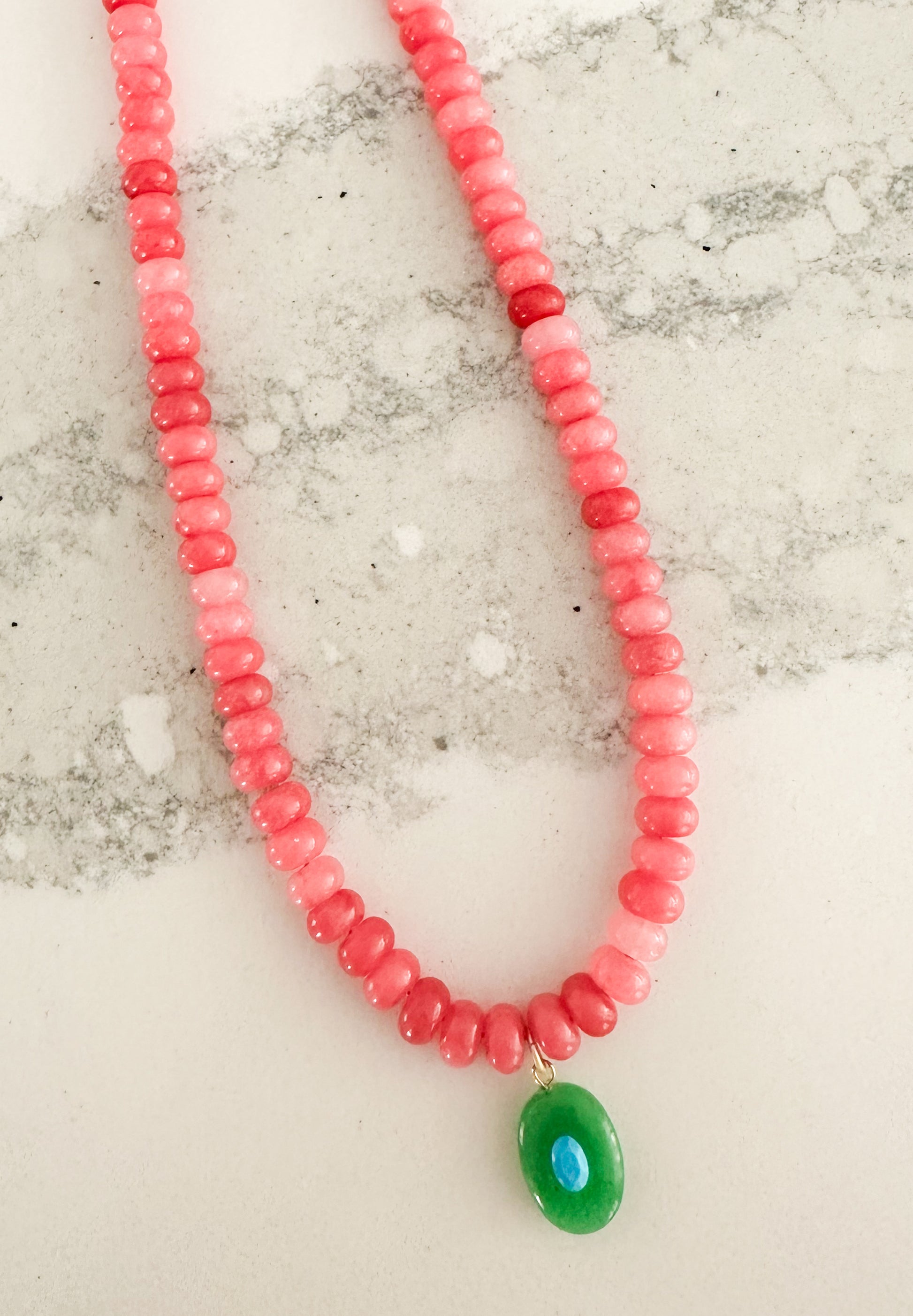 A beaded necklace made of small pink and red beads with a green oval pendant, displayed on a light marble-like surface. The Sawyer Necklace