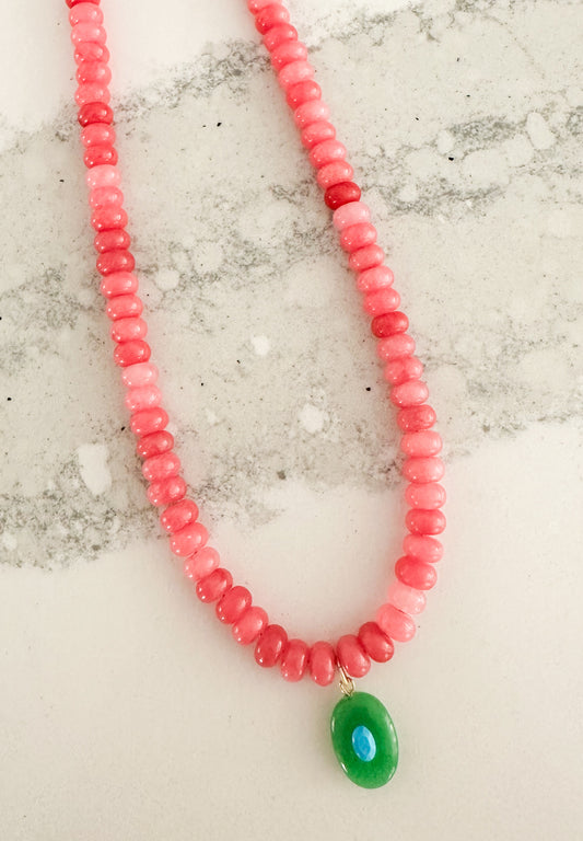 A beaded necklace made of small pink and red beads with a green oval pendant, displayed on a light marble-like surface. The Sawyer Necklace