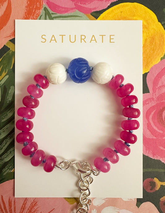 A beaded bracelet displayed on a white Saturate card, featuring bright pink round beads, two white carved beads, and one blue faceted bead, with a silver clasp. The card is set against a colorful floral background.