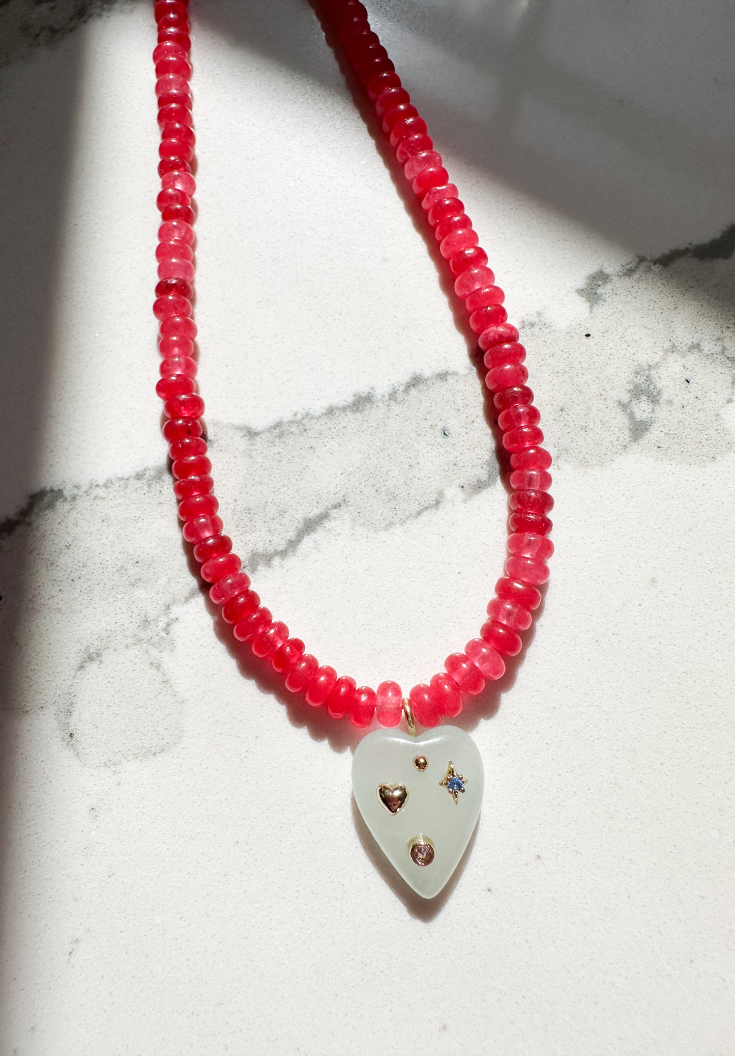 Ella necklace with a white heart-shaped pendant decorated with small gold and blue stones, displayed on a white marble surface with dramatic shadows.