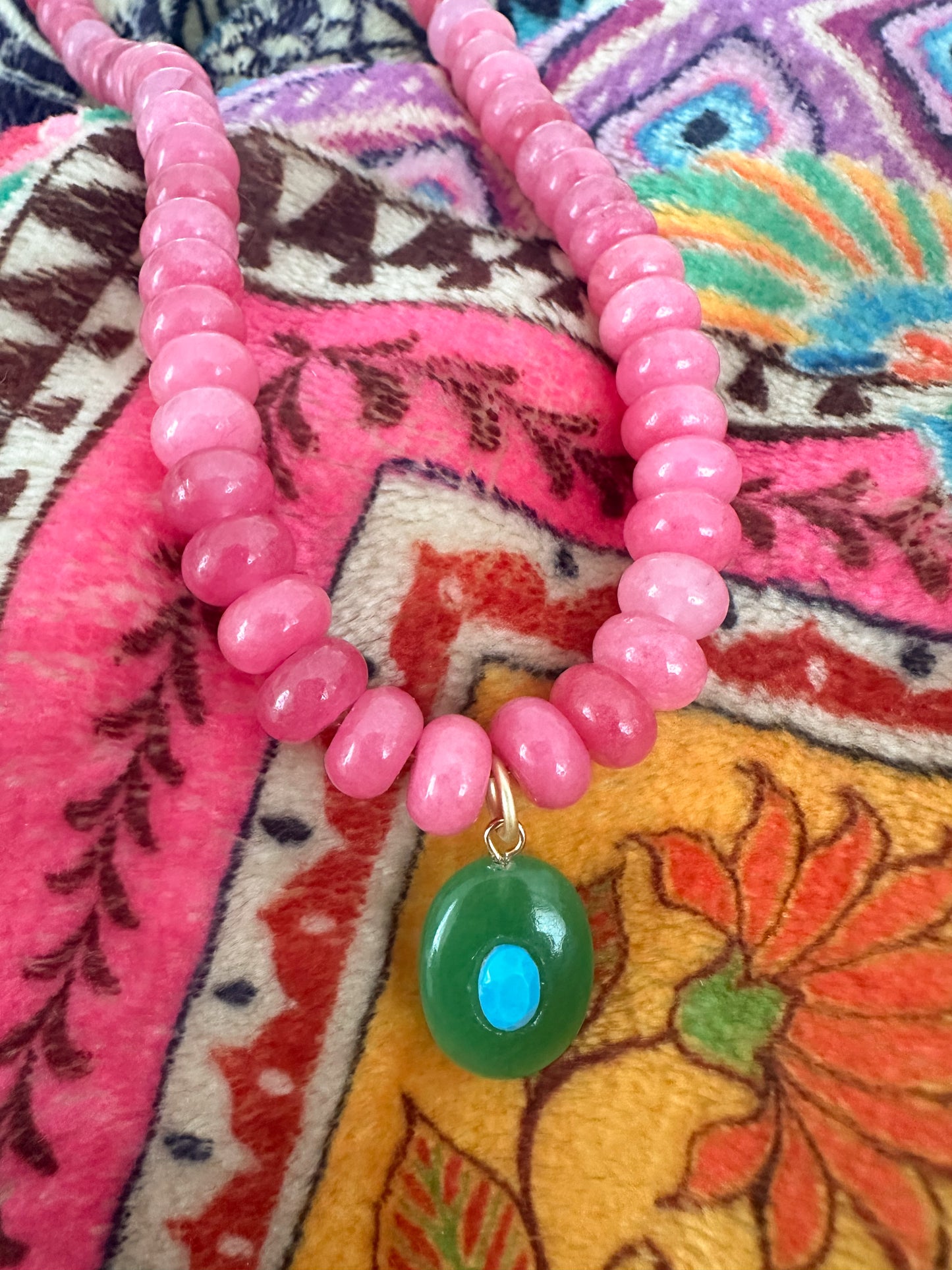 A pink beaded necklace with a green oval pendant featuring a blue center, displayed on a colorful patterned textile background.