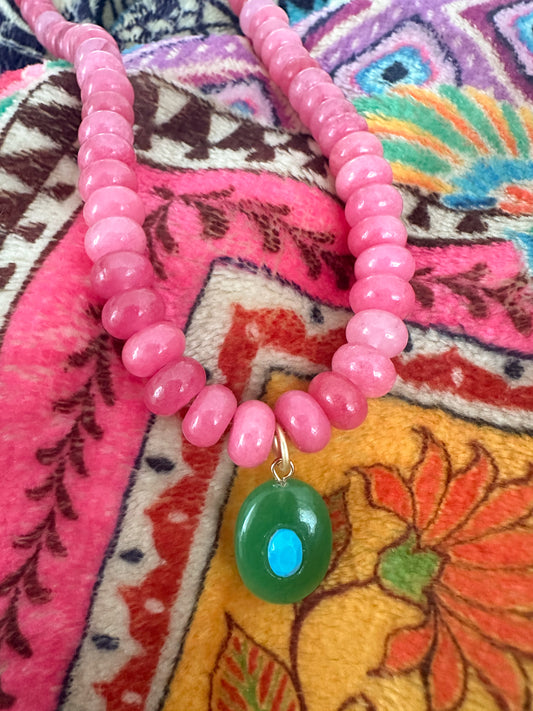 A pink beaded necklace with a green oval pendant featuring a blue center, displayed on a colorful patterned textile background.