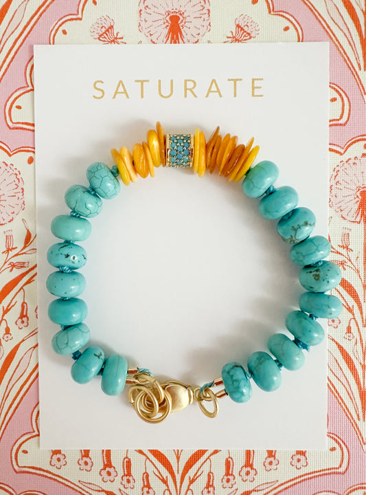 A turquoise and orange beaded bracelet with a gold clasp, displayed on a white Saturate brand card against a pink and orange patterned background.